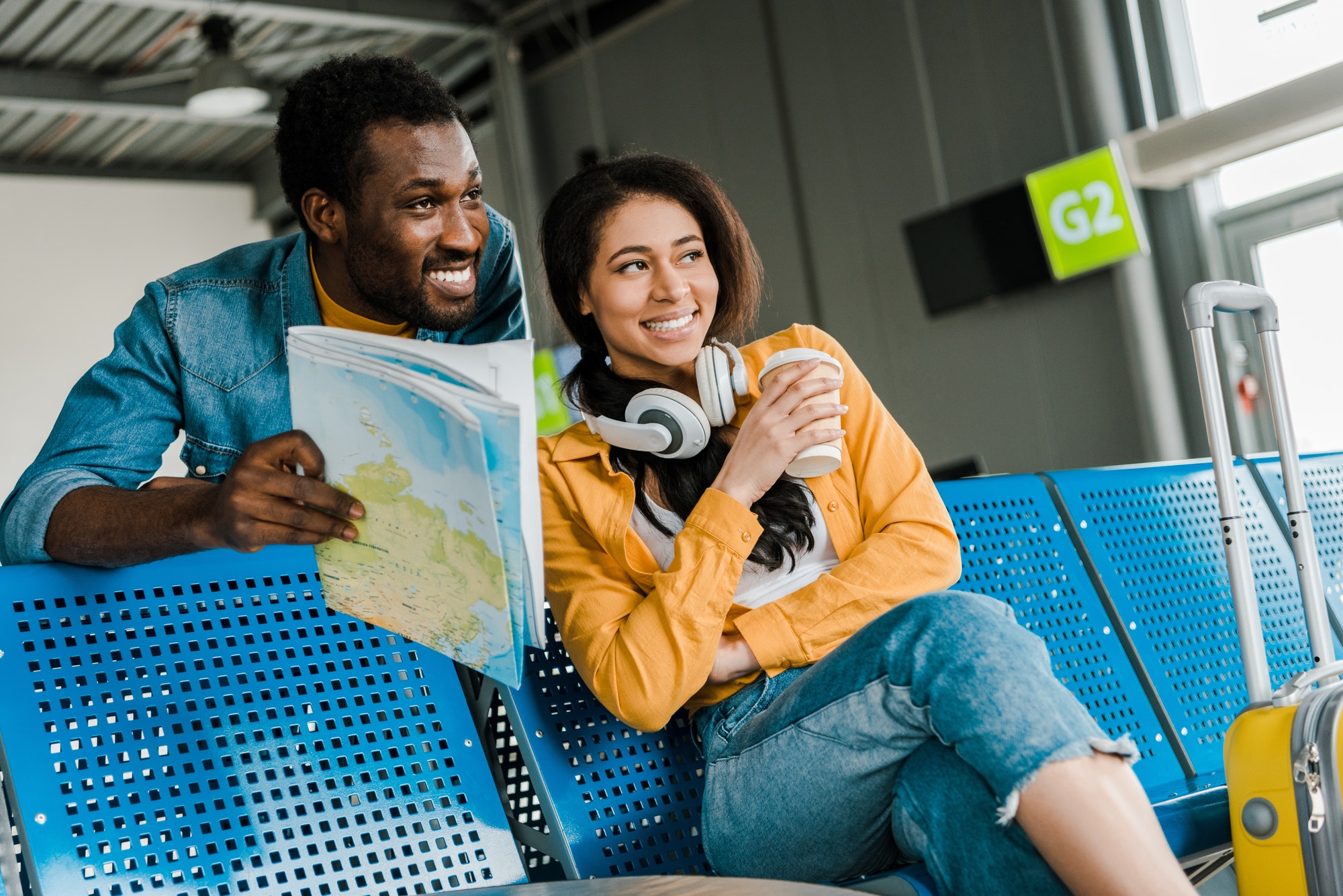 smiling african american couple sitting with map in departure lounge in airport and looking away
